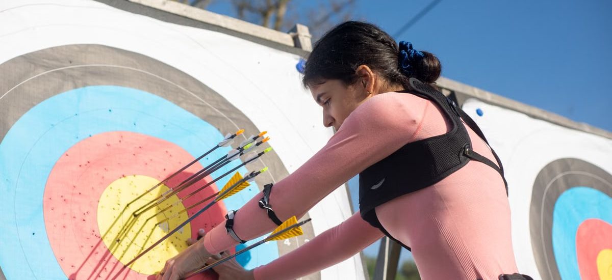 A female archer adjusts arrows on a target during a sunny day, showcasing precise archery skills.